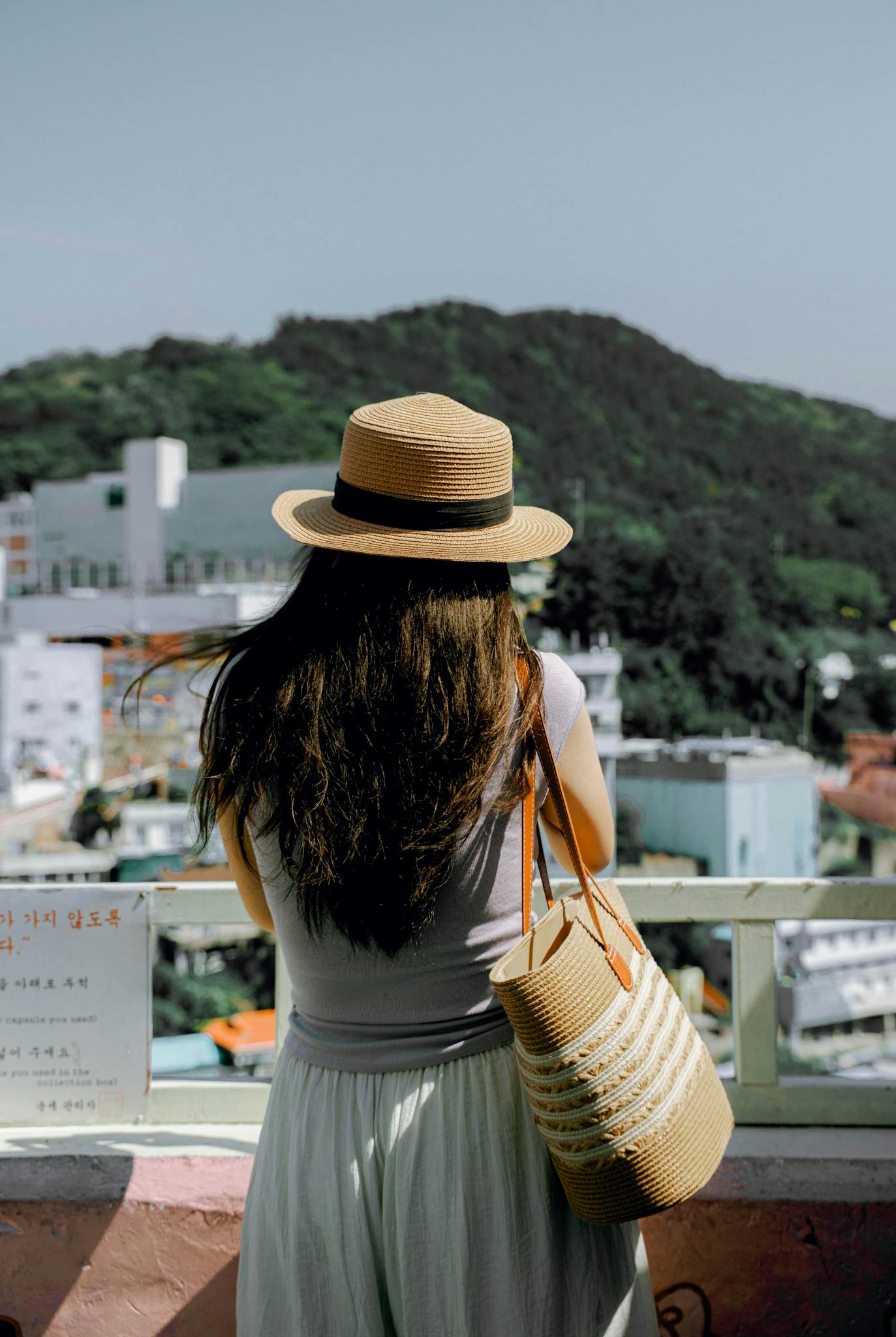 A woman in a straw hat overlooks a picturesque cityscape and hills in South Korea.